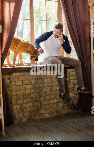 Vista del giovane uomo giocando con il cane e parlando al telefono. Foto Stock