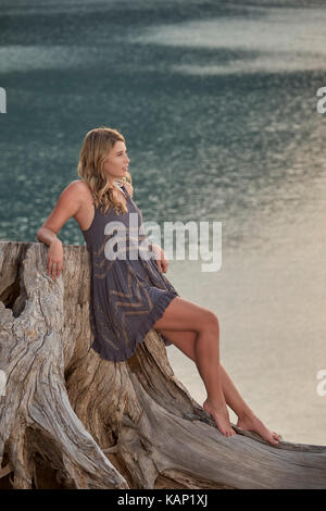 Una giovane donna relax su un ceppo di albero accanto ad un piccolo lago di montagna Foto Stock