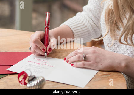 Giovane donna scrivere cartoline di Natale con unghie rosse, una penna rossa e decorazioni di vacanza su una tavola di legno Foto Stock