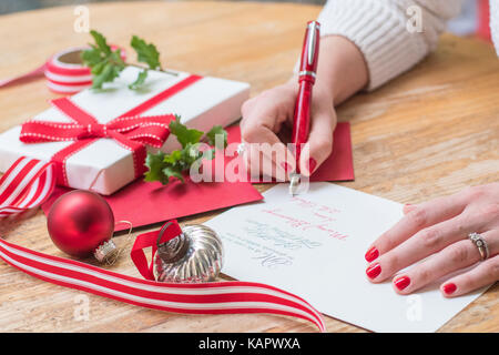 Giovane donna scrivere cartoline di Natale con unghie rosse, una penna rossa e decorazioni di vacanza su una tavola di legno Foto Stock