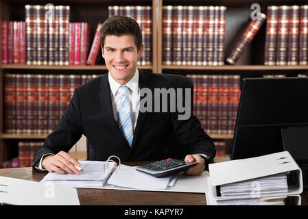 Ritratto di fiducia avvocato utilizzando la calcolatrice mentre la scrittura su documenti alla scrivania in ufficio Foto Stock