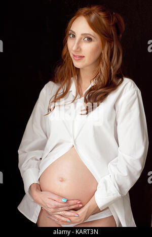 I capelli rossi giovane donna incinta su sfondo nero che indossa in camicia bianca. lei si mette le mani sulla pancia per sentire il suo bambino. Foto Stock