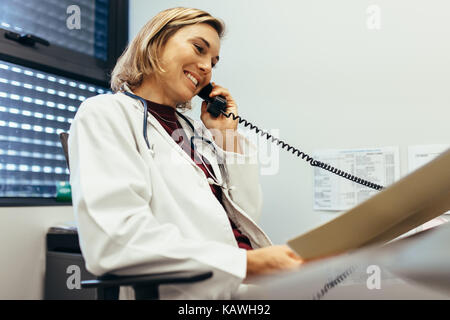 Medico donna la lettura dei rapporti medici e parlando al telefono alla clinica. Medico a lavorare alla sua scrivania in ufficio. Foto Stock