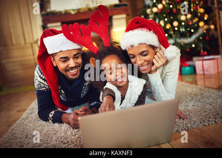 Bella la Condivisione in famiglia laptop vicino all'albero di natale in una sera d'inverno, godendo la calda atmosfera natalizia nel loro salotto e indossando un ettaro Foto Stock