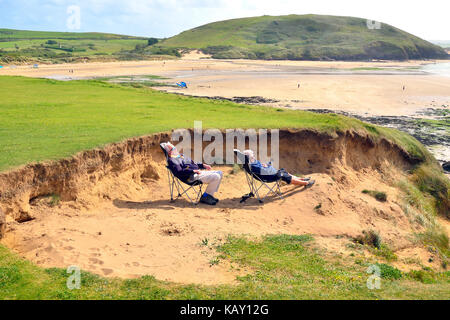 Uomo maturo e la donna riparo dal vento nelle dune di sabbia mentre crogiolandovi sotto i raggi del sole all'Daymer Bay, Cornwall, England, Regno Unito Foto Stock