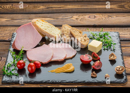 Pezzi di cold-bollite, pane, burro, pomodori, erbe, noci, la senape. Ingredienti per la prima colazione sulla pietra nera scheda sopra lo sfondo di legno Foto Stock