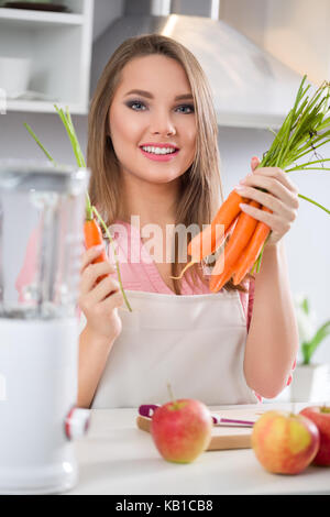 Giovane donna allegra in cucina azienda carote, pronti a rendere frullati di frutta con frullatore Foto Stock