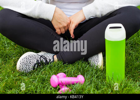 Close up femminile atletica seduta con le gambe incrociate su erba con manubri e una bottiglia di acqua Foto Stock