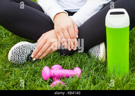 Close up femminile atletica seduta con le gambe incrociate su erba con manubri e una bottiglia di acqua Foto Stock