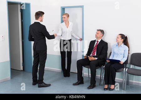 Imprenditrice stringono le mani con l'uomo di fronte a persone in attesa per il colloquio di lavoro in ufficio Foto Stock