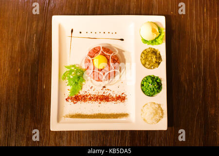 Gustoso piatto di carne cruda, tuorlo d'uovo, verdi e spezie immessi sul bianco piastra rettangolare. La vista di cui sopra. Foto Stock