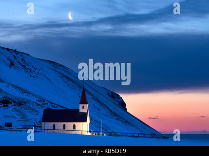 Chiesa illuminata all'alba contro montagne coperte di neve, in inverno o in prossimità di vik, sud Islanda, regioni polari Foto Stock