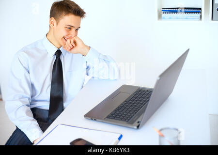 Ritratto di giovane imprenditore ridere guardando lo schermo del laptop al suo luogo di lavoro in ufficio luminoso Foto Stock