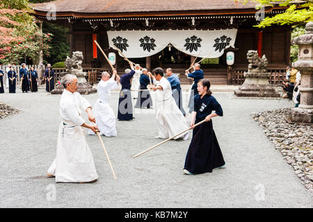 Giappone, Osaka, Tada. Master spadaccino samurai vestito di bianco a combattere con la donna in nero, utilizzando bokken o bokuto, spade di legno nel santuario terreno. Foto Stock