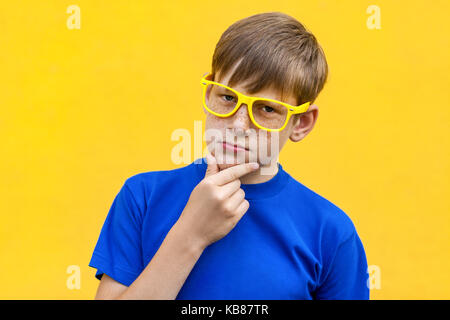 Grave freckled boy guardando la telecamera, toccando il mento e pensare. parete gialla, studio shot Foto Stock