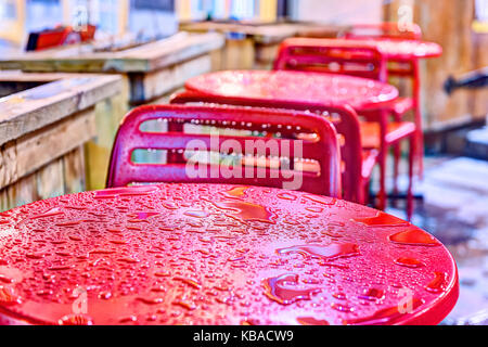 Wet red tavolo, sedie closeup fuori del ristorante area salotto dal marciapiede durante forti piogge durante la notte con una pozza, gocce, goccioline di acqua Foto Stock