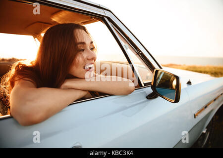 Lato Vista ravvicinata della donna sorridente in occhiali da sole seduti all'interno di una vettura e guardando fuori della finestra Foto Stock