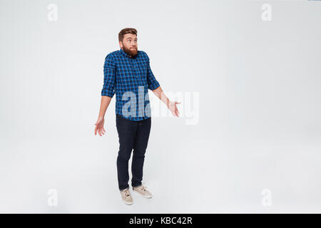 A piena lunghezza Ritratto di un confuso giovane uomo barbuto in piedi e scrollare le spalle isolate su sfondo bianco Foto Stock