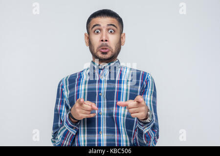 L'uomo seriosly, indossa una maglietta blu, bocche di apertura ampiamente, dopo aver stupito sconvolto guarda, puntare il dito alla telecamera. isolato studio shot su grigio b Foto Stock