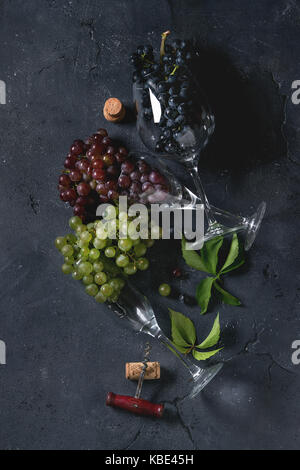 Varietà di tre tipo fresche Uve mature blu scuro, rosso e verde in posa diversi bicchieri di vino con la vecchia struttura di cavatappi e foglie verdi sul nero textu Foto Stock