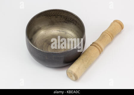 Un Singing Bowl, fatta di metallo, e un bastone di legno che giace accanto ad essa, su una superficie bianca. Foto Stock