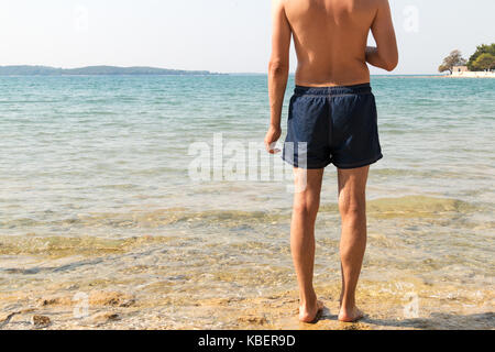 Modello maschile di spalle guardando il mare. Acqua chiara, con sole splendente e protezione solare per la scottatura. l uomo in pantaloncini blu ottenere pronto ad andare a nuotare in Foto Stock