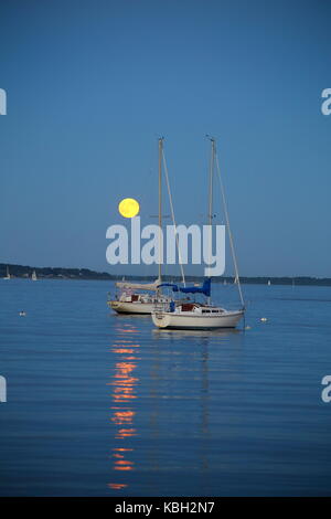 Luna piena di Harvest che sorge sulla baia di Narragansett, in Rhode Island, nel New England meridionale, in una bella serata con barche a vela in acqua Foto Stock