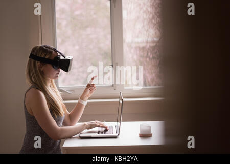 Vista laterale della donna con la realtà virtuale auricolare tramite computer portatile dalla finestra a casa Foto Stock