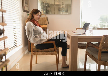 Donna incinta a leggere un libro mentre è seduto su una sedia Foto Stock