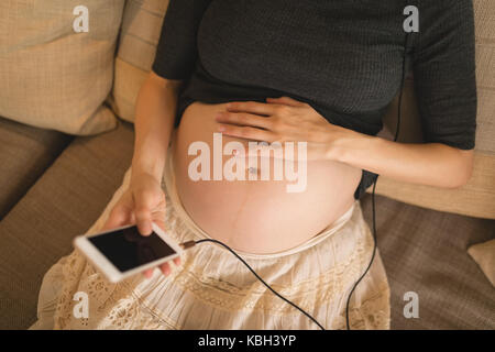 Donna incinta usando il telefono cellulare mentre si tocca il suo ventre a casa Foto Stock