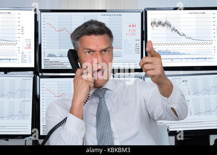 Maschio arrabbiato stock trader gridando mentre si utilizza il telefono con schermi di computer in background Foto Stock