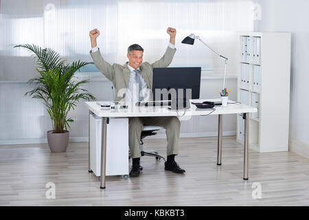 Imprenditore di successo con le braccia sollevate celebrando la vittoria al computer scrivania in ufficio Foto Stock