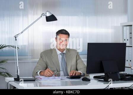 Ritratto di sorridere ragioniere utilizzando la calcolatrice mentre la scrittura su documenti alla scrivania in ufficio Foto Stock