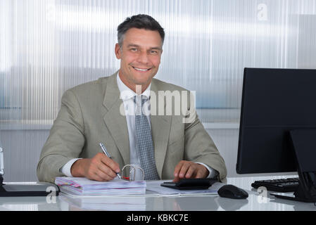 Ritratto di fiducioso ragioniere utilizzando la calcolatrice mentre la scrittura su documenti alla scrivania in ufficio Foto Stock