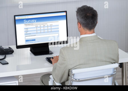 Vista posteriore di imprenditore riempiendo il modulo del sondaggio sul computer alla scrivania in ufficio Foto Stock