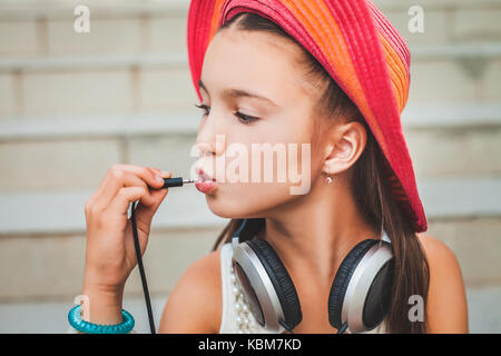 Funny Girl mette il tappo nella sua bocca. il bambino in un Red Hat, camicia bianca e con le cuffie grigio sullo sfondo di scale Foto Stock