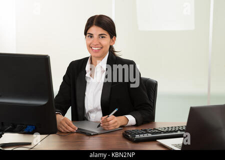 Felice giovani designer femmina con tavoletta grafica seduto alla scrivania Foto Stock
