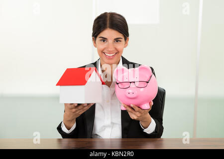 Felice giovane imprenditrice azienda modello di casa e piggybank in mani Foto Stock