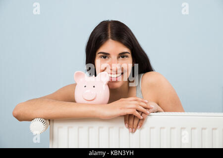 Giovane donna felice con Piggybank sul radiatore a casa Foto Stock