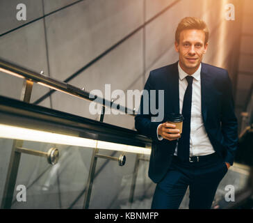 Sorridente giovane imprenditore in un vestito a bere un caffè e salire una scala mobile in una stazione della metropolitana durante i suoi spostamenti di mattina