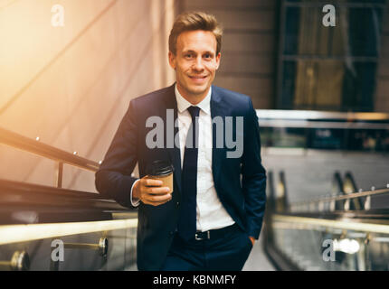 Fiducioso giovane imprenditore sorridere mentre bere un caffè e salire una scala mobile in una stazione della metropolitana durante i suoi spostamenti di mattina