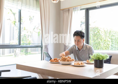 Giovane uomo asiatico di mangiare pollo fritto nel salotto di casa contemporanea per lo stile di vita moderno concetto Foto Stock