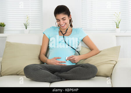 Ritratto di felice donna incinta ascoltando baby heartbeat attraverso uno stetoscopio sul divano di casa Foto Stock