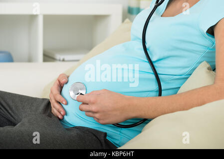 Sezione mediana della donna incinta tenendo uno stetoscopio su stomaco mentre è seduto sul divano di casa Foto Stock