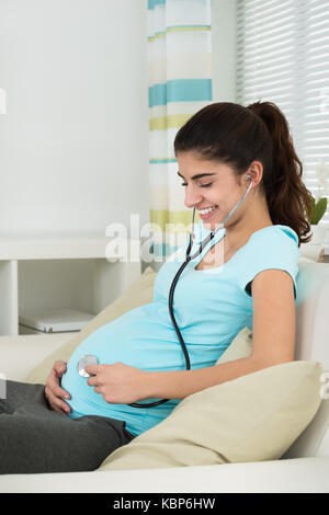 Ritratto di felice donna incinta ascoltando baby heartbeat attraverso uno stetoscopio sul divano di casa Foto Stock