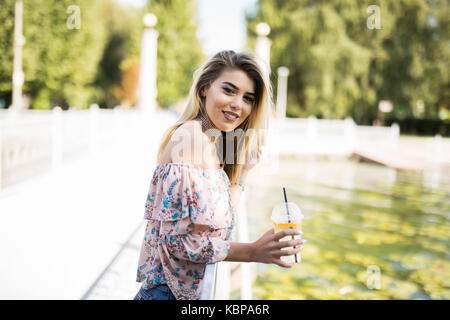 Felice ragazza bionda seduta al parco, bere una bottiglia di freddo succo verde mentre sorridente nella distanza. Foto Stock