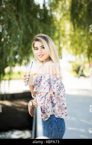 Felice ragazza bionda seduta al parco, bere una bottiglia di freddo succo verde mentre sorridente nella distanza. Foto Stock