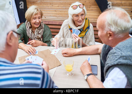 Due coppie senior indossando vestiti caldi carta da gioco con ciascun altro mentre sono riuniti presso accogliente piccolo patio Foto Stock