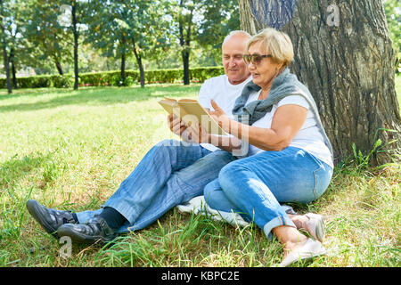 A piena lunghezza Ritratto di amorevole coppia senior indossano jeans e T-shirt bianche, seduto sotto agli alberi e leggendo la loro avventura preferiti storia insieme, Foto Stock