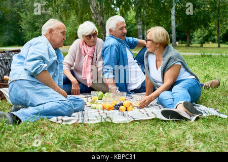 Due coppie senior godendo di ogni altri società pur avendo picnic al parco verde, essi seduti sul plaid Coperte con fresca frutta succosa Foto Stock